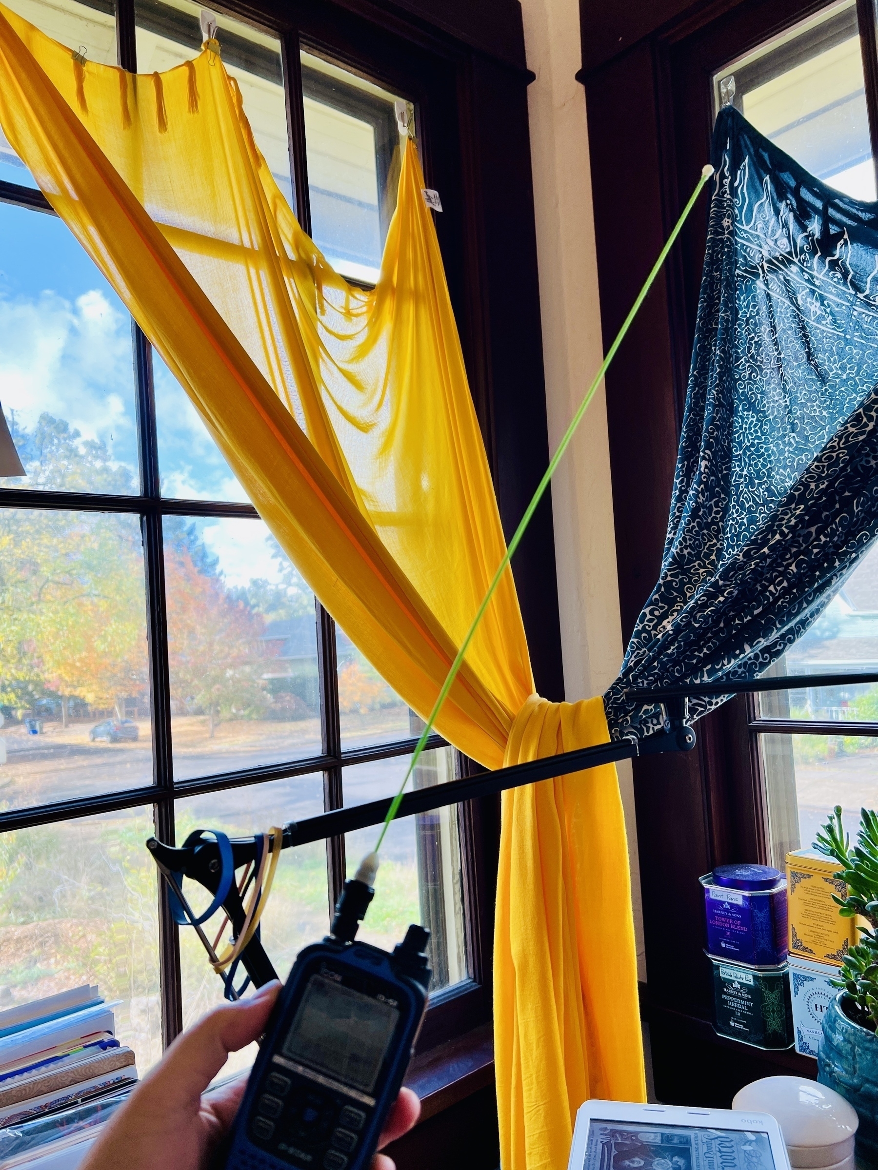 Ham radio handheld being held in the hand, with a lime green whip antenna. Yellow and blue curtains visible in the background, and the edges of a messy workspace. 